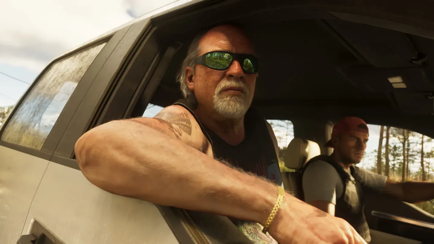 Hombre mayor con perilla blanca, gafas de sol envolventes y camiseta de tirantes, con el brazo tatuado apoyado en la ventanilla de una furgoneta. En el asiento de al lado va un hombre joven con gorra.