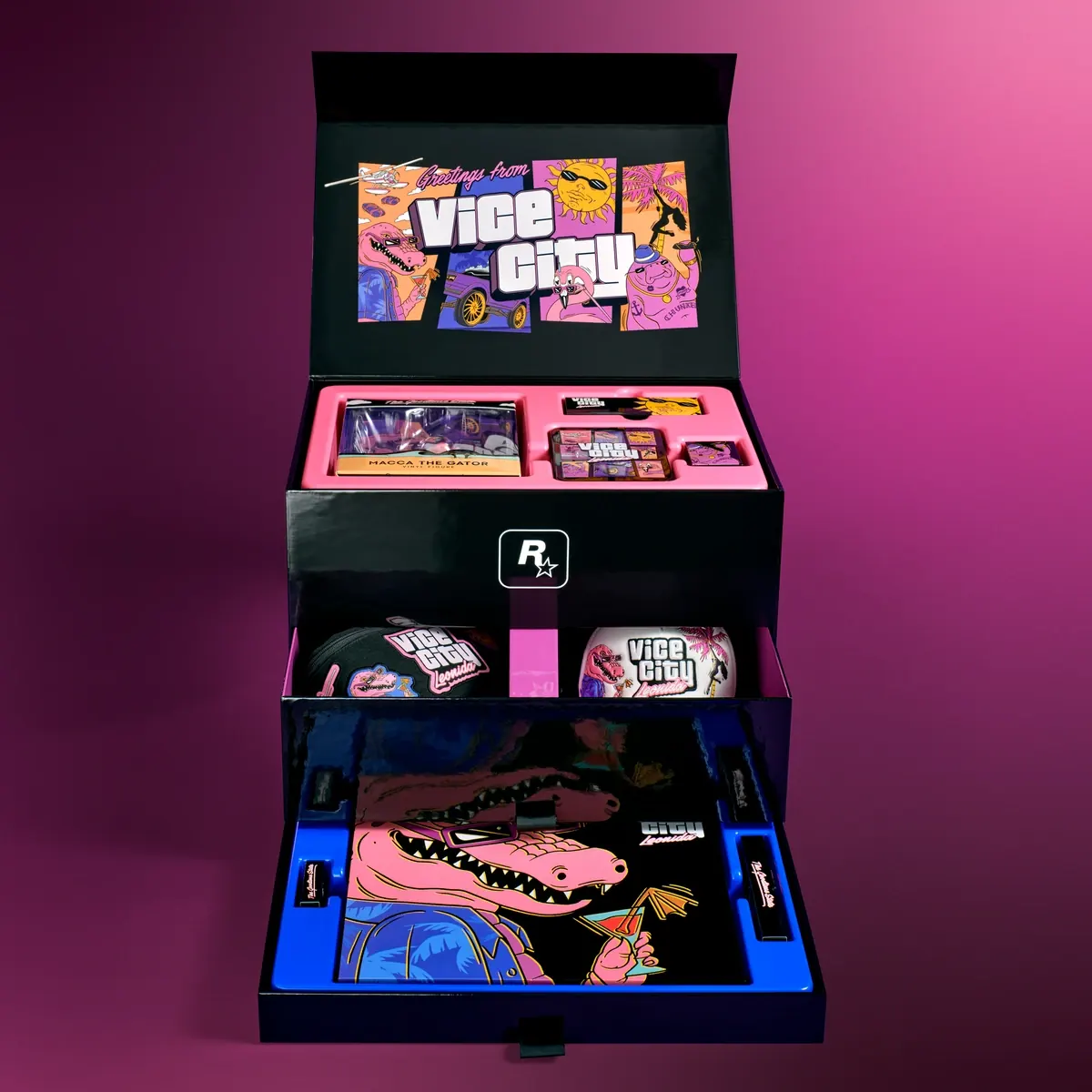 La misma caja, abierta. Por dentro de la tapa, una ilustración de «Greetings from Vice City» con personajes de dibujo; debajo, una bandeja rosa con la figura de Macca en su caja y otros objetos, y dos cajones abiertos con más piezas.