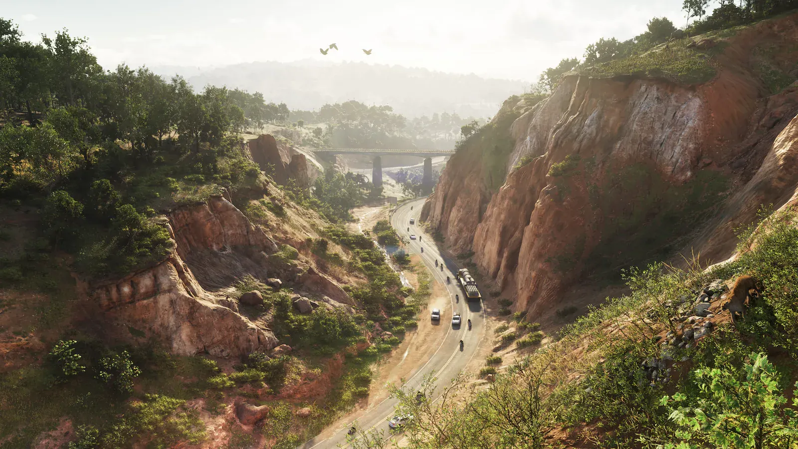 Carretera que serpentea por un cañón de roca rojiza, con coches y motos circulando y un puente al fondo entre la bruma.