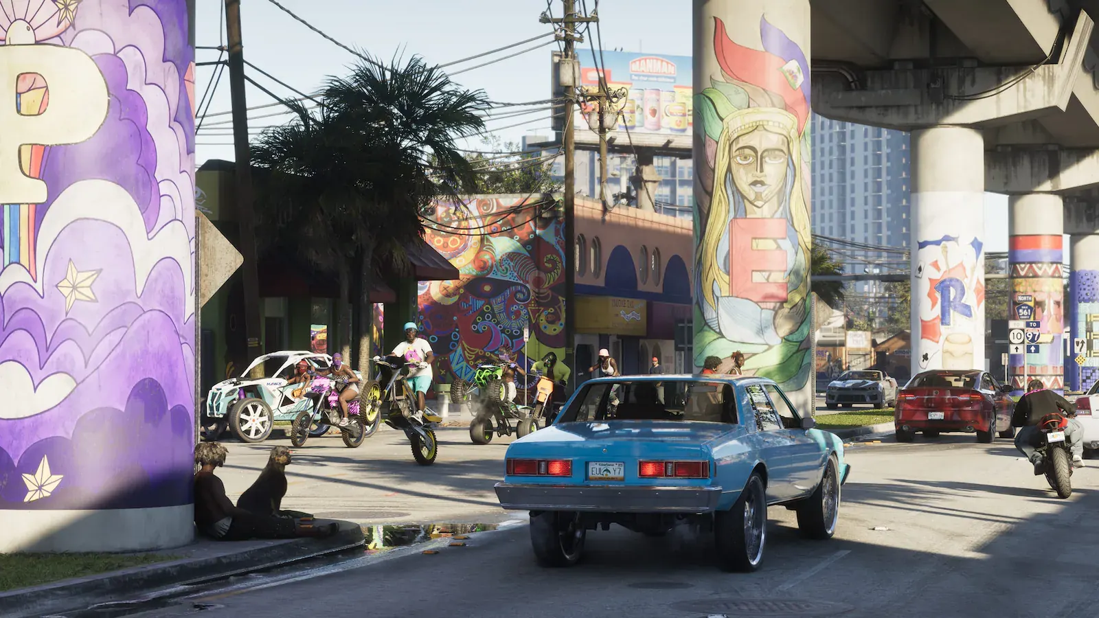 Calle bajo una autopista elevada cuyos pilares están pintados con murales de colores. Un sedán clásico azul circula y varias personas van en motos de cross y quads.
